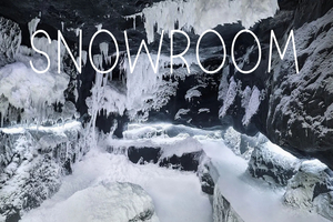 Wnętrze lodowej jaskini lub pomieszczenia typu snowroom z czarnymi skałami pokrytymi grubą warstwą śniegu i wiszącymi soplami lodu. Na górze obrazu widnieje duży, biały napis „SNOWROOM”.